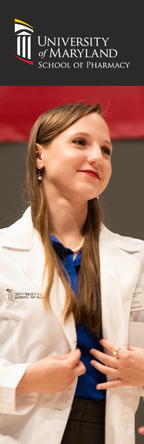 Person wearing a white lab coat stands indoors in front of a University of Maryland School of Pharmacy backdrop. The lab coat features the School of Pharmacy logo on the chest, and the person wears a blue top underneath while adjusting the front of the coat. The background includes a red and dark gray wall with the University of Maryland School of Pharmacy name and logo displayed above.