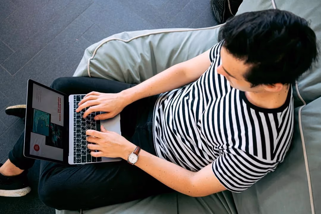 Person sitzt mit Laptop auf dem Schoß auf einem Sofa und tippt, von oben fotografiert