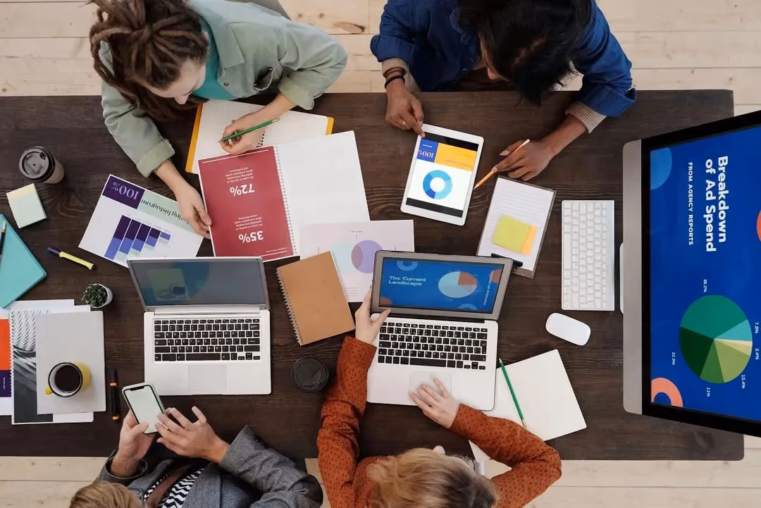 Vier Menschen sitzen um einen Tisch mit Laptops, Tablets und ausgedruckten Diagrammen, von oben fotografiert