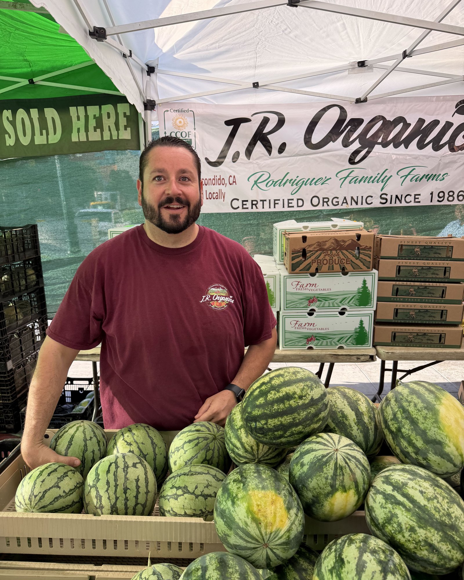 Michael Clark behind crates of watermelons at the JR Organics market stand, under a Rodriguez Family Farms banner reading Certified Organic Since 1986