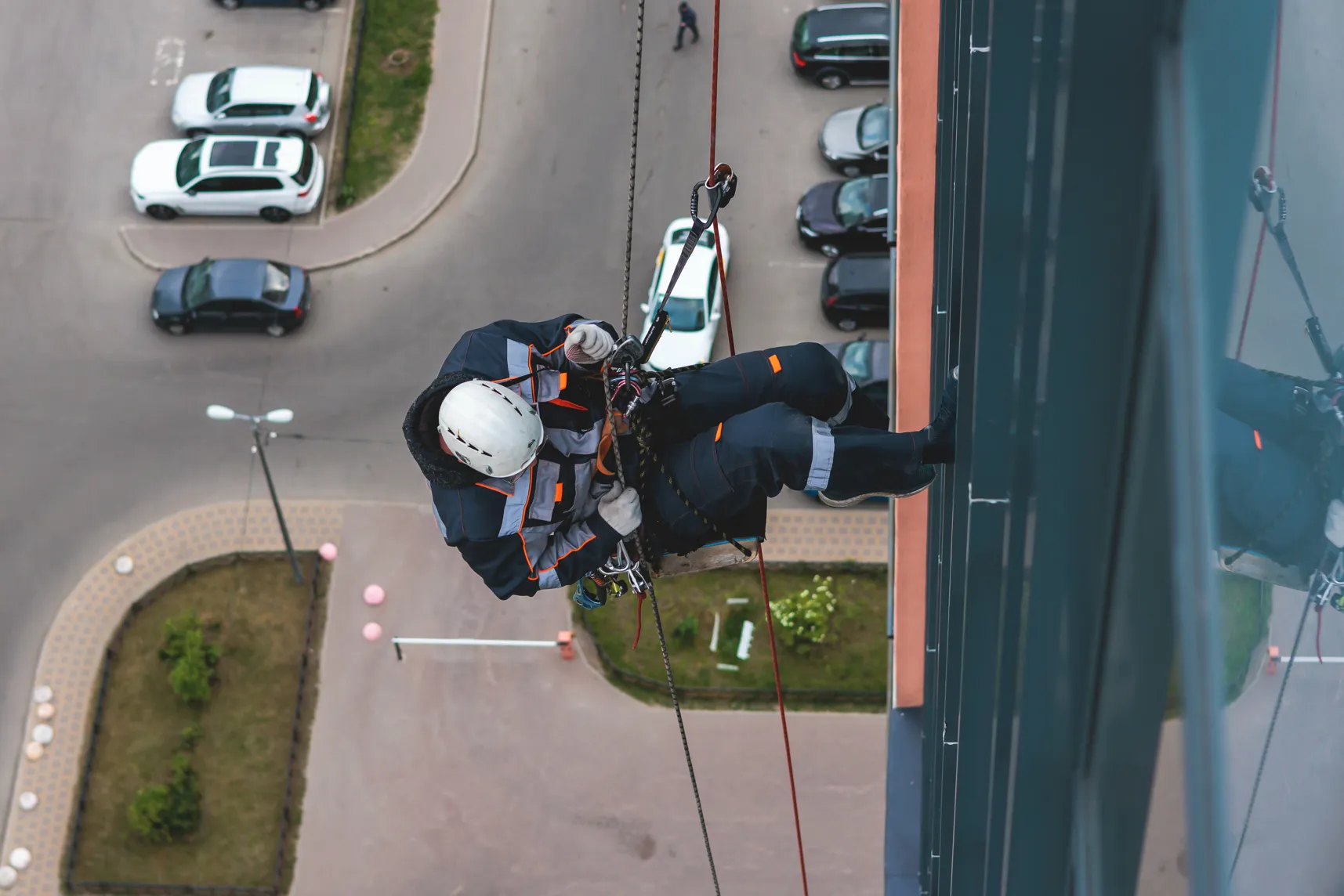 Window cleaner rappelling down building exterior with parked cars below