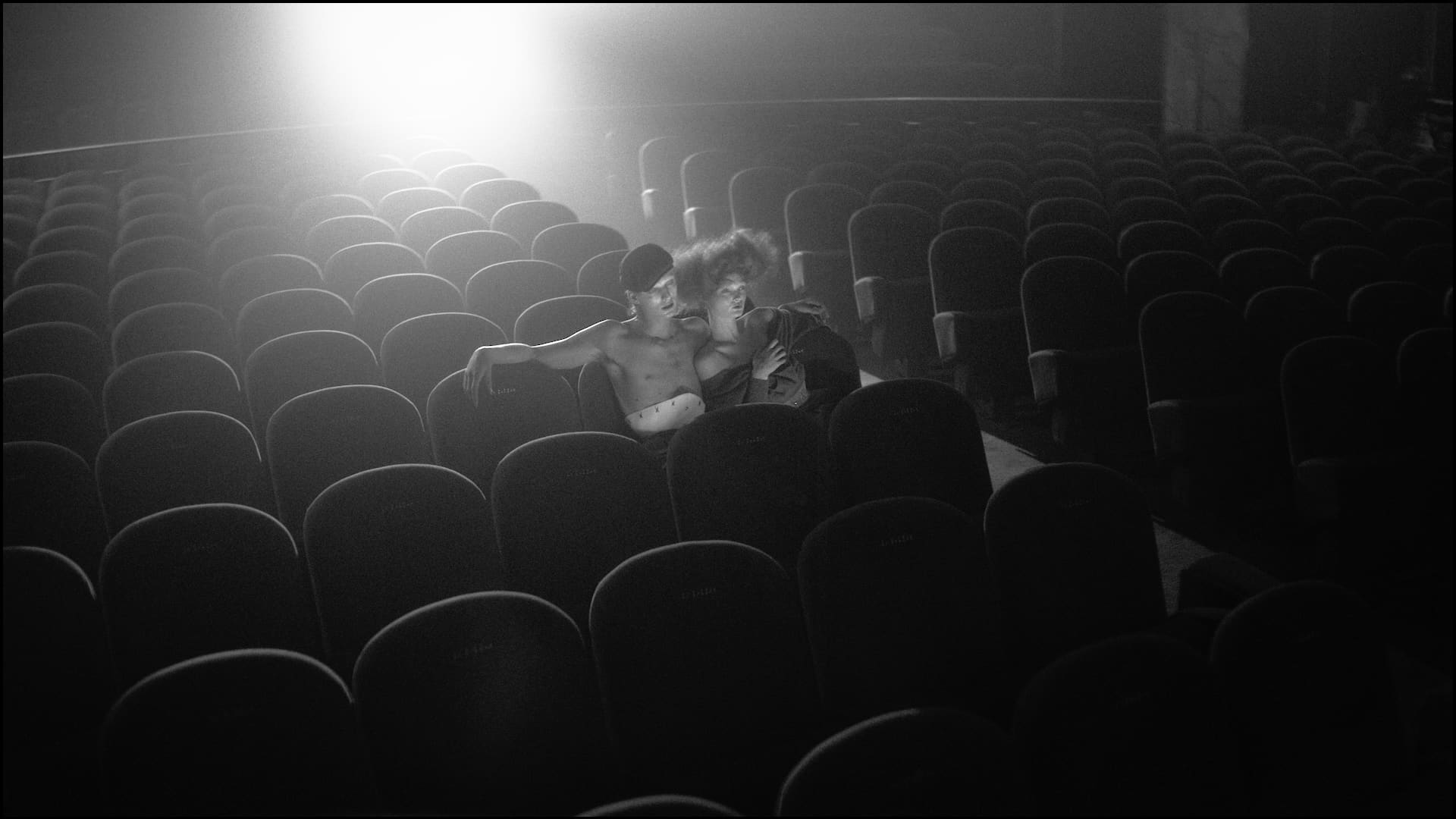 Nighthawk: two figures seated in an empty cinema, surrounded by rows of seats and projector light, in black and white.