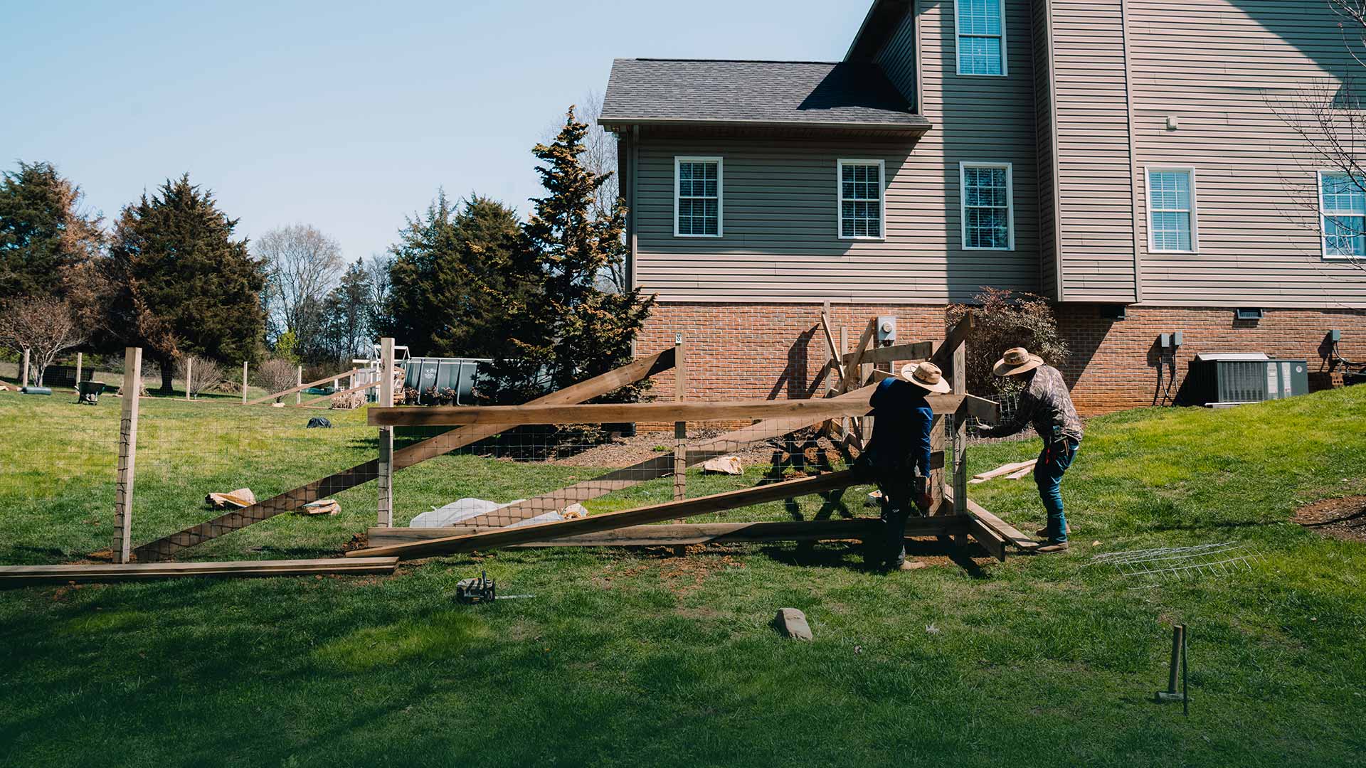 Two-person crew handling long wooden rails for a new fence on a sloping lawn beside a house