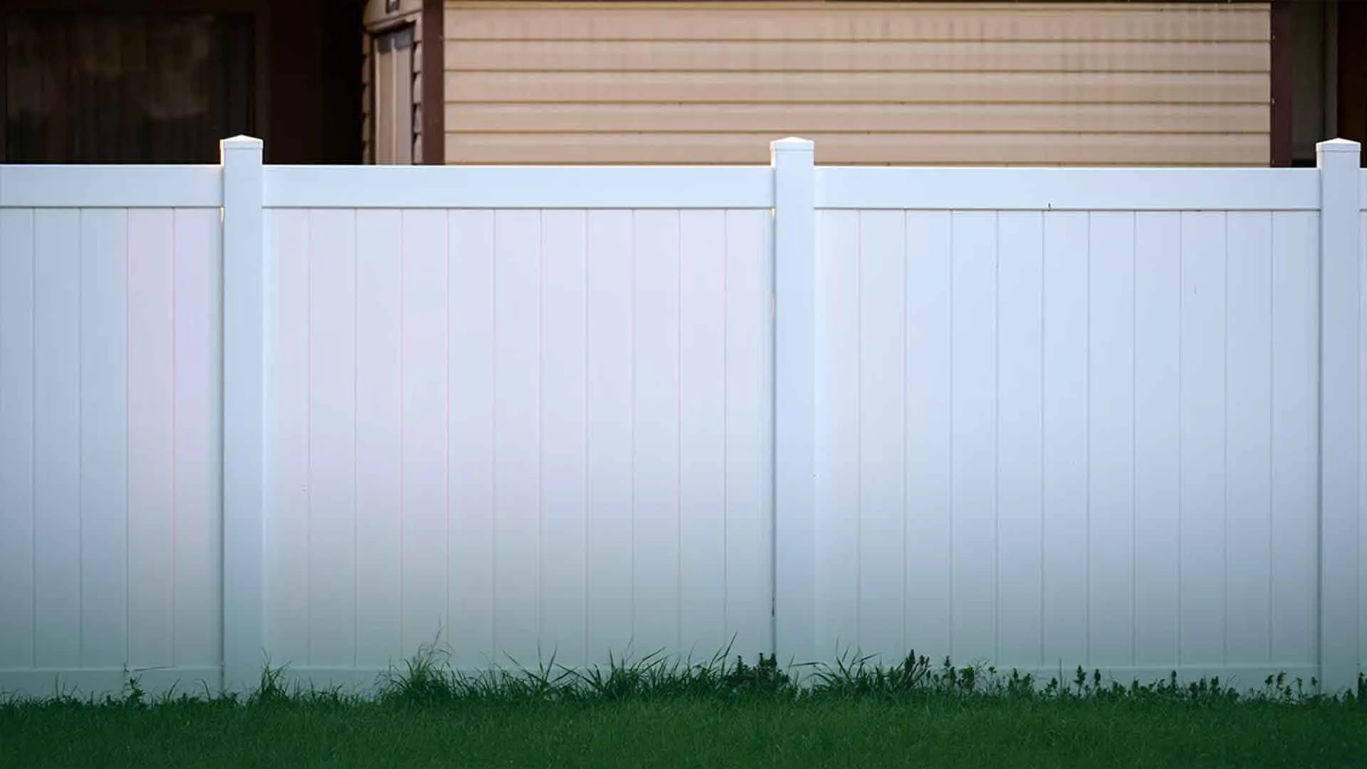 Solid white vinyl privacy fence panels standing between a beige-sided house and the lawn