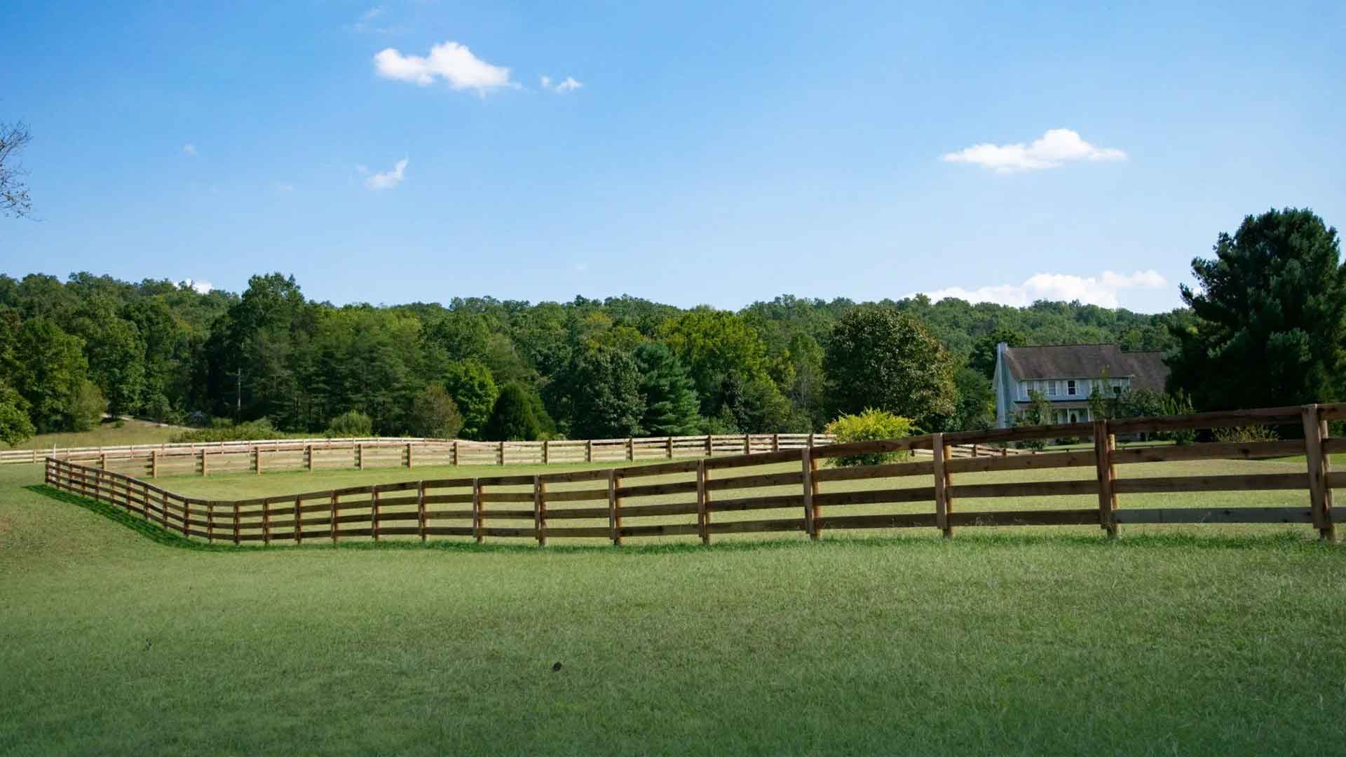 Wood board fence turning a corner around a wide green pasture below a wooded ridge