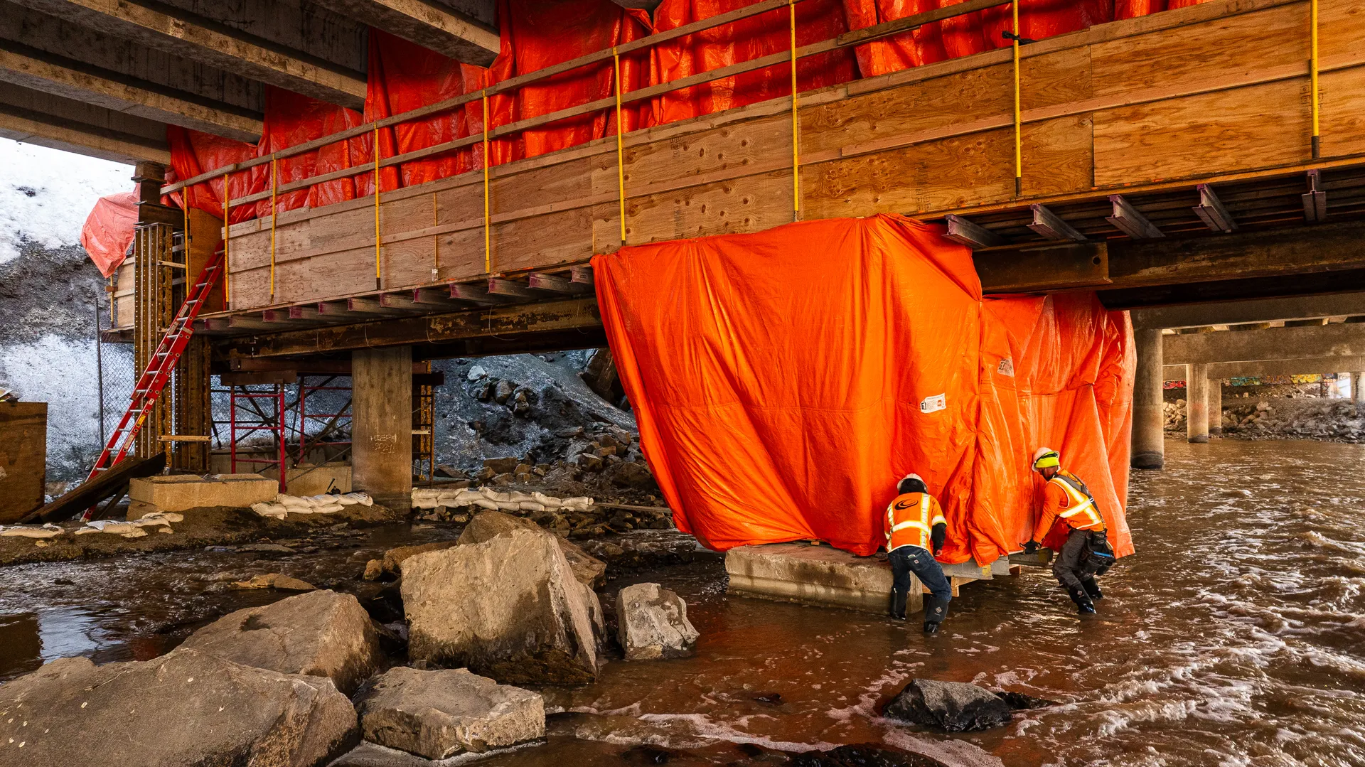 Two Gerber crew members work under a bridge deck beside a creek, beneath orange containment tarps, in Ogden Canyon.