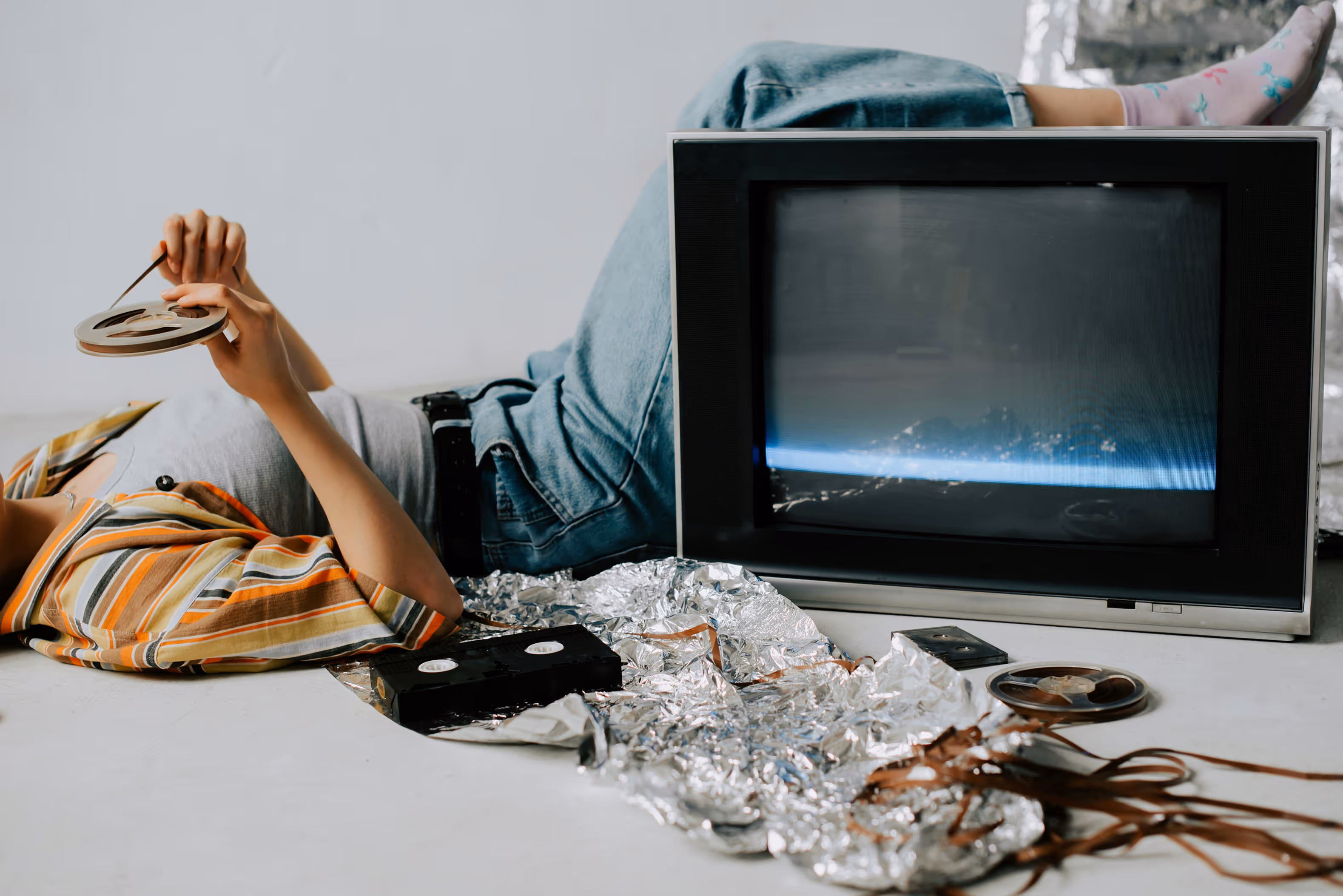 Une personne allongée au sol tient une bobine de film à côté d’une vieille télévision et de cassettes VHS