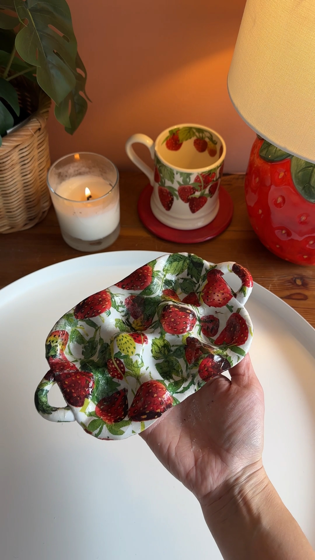 a handmade clay tray covered in a strawberry pattern, held up beside a candle and a strawberry lamp