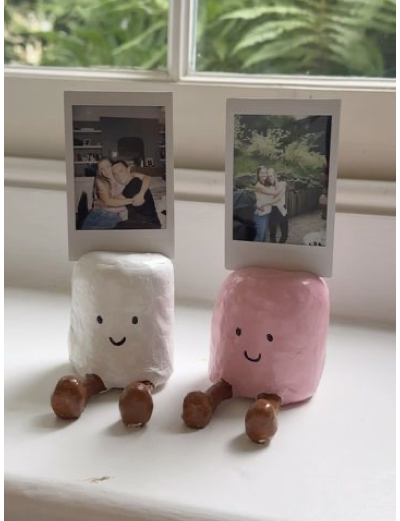 two smiling marshmallow-shaped clay characters with little feet, each holding up a polaroid photo on a windowsill
