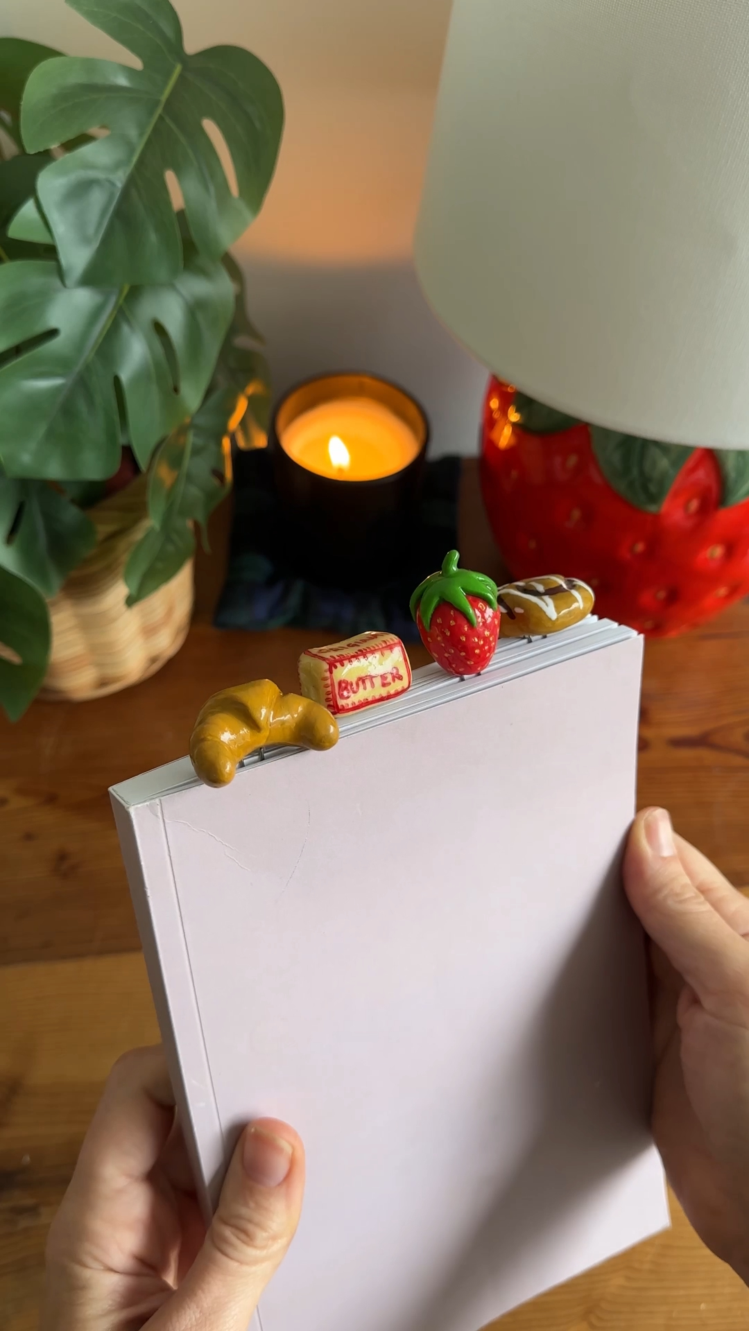 four tiny clay bookmarks shaped like a croissant, a pat of butter, a strawberry and an iced bun, clipped to the top of a book