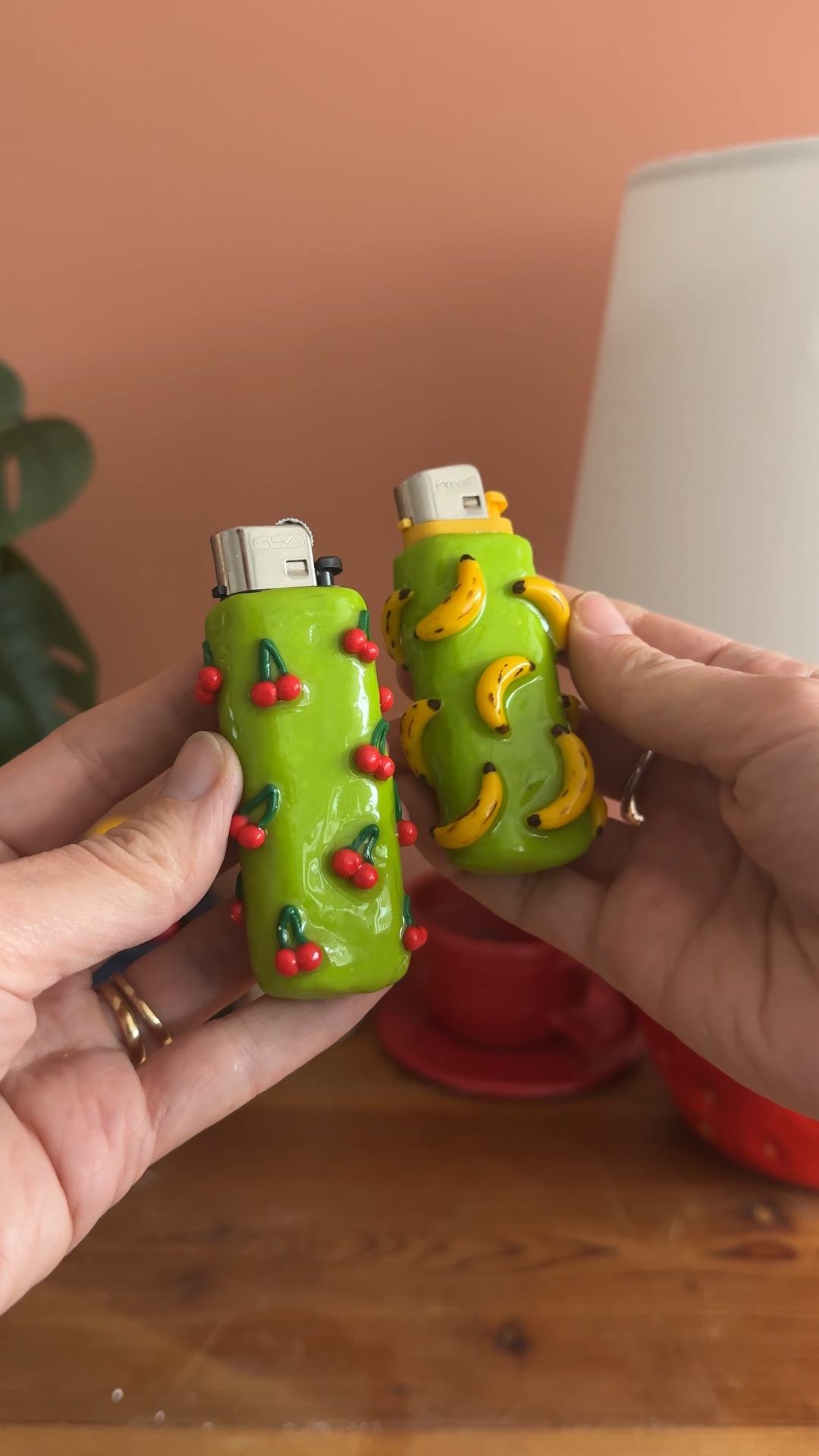 two lighters covered in green clay, one decorated with tiny red cherries and one with tiny yellow bananas