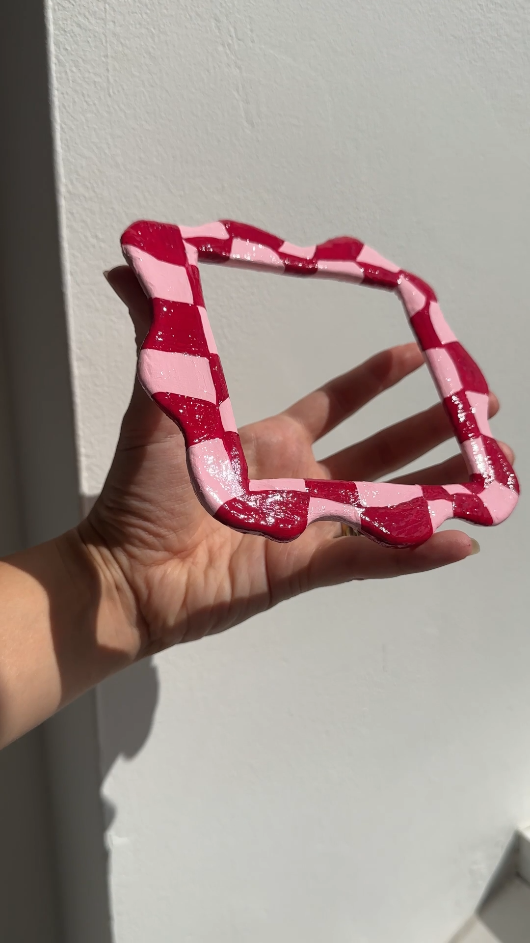 a square clay frame painted in red and pink checkerboard, held up against a white wall