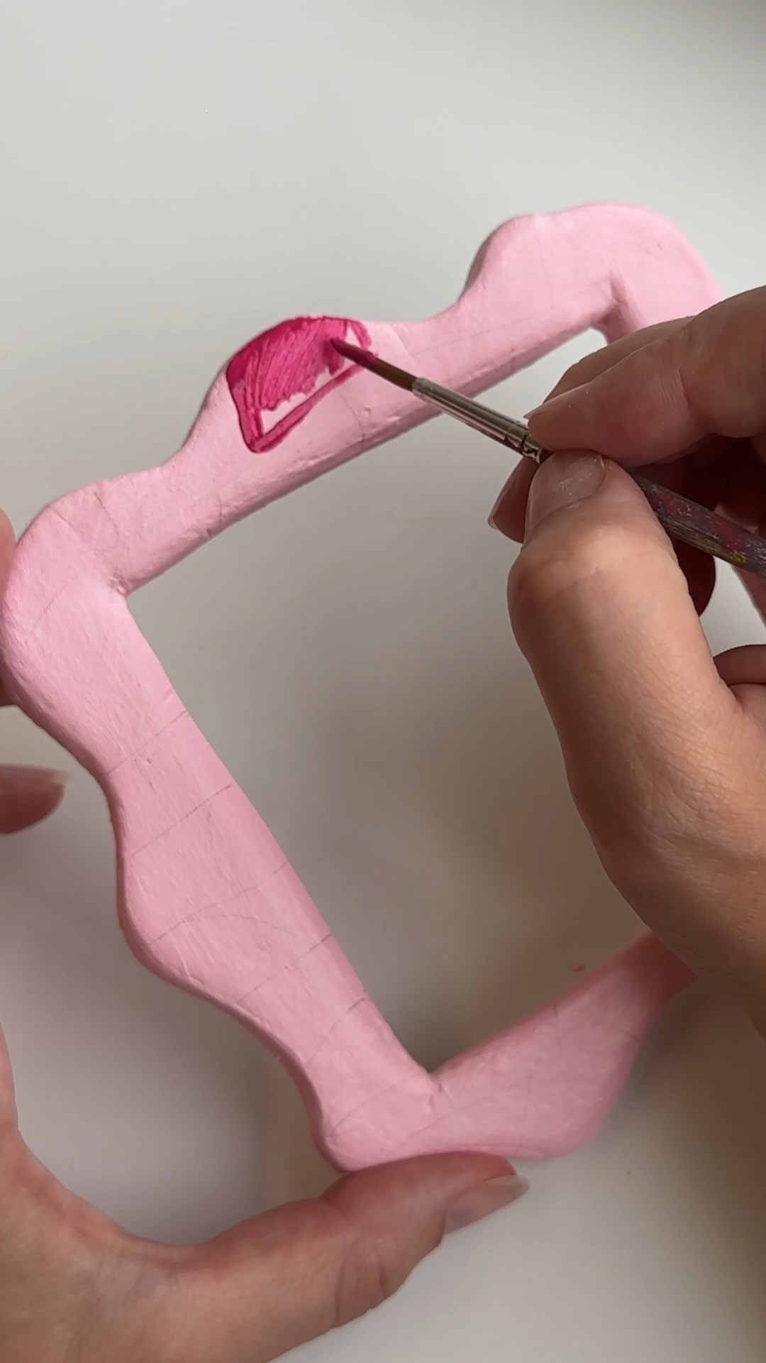 hands painting a pink clay frame, filling in one checkerboard square at a time with a fine brush
