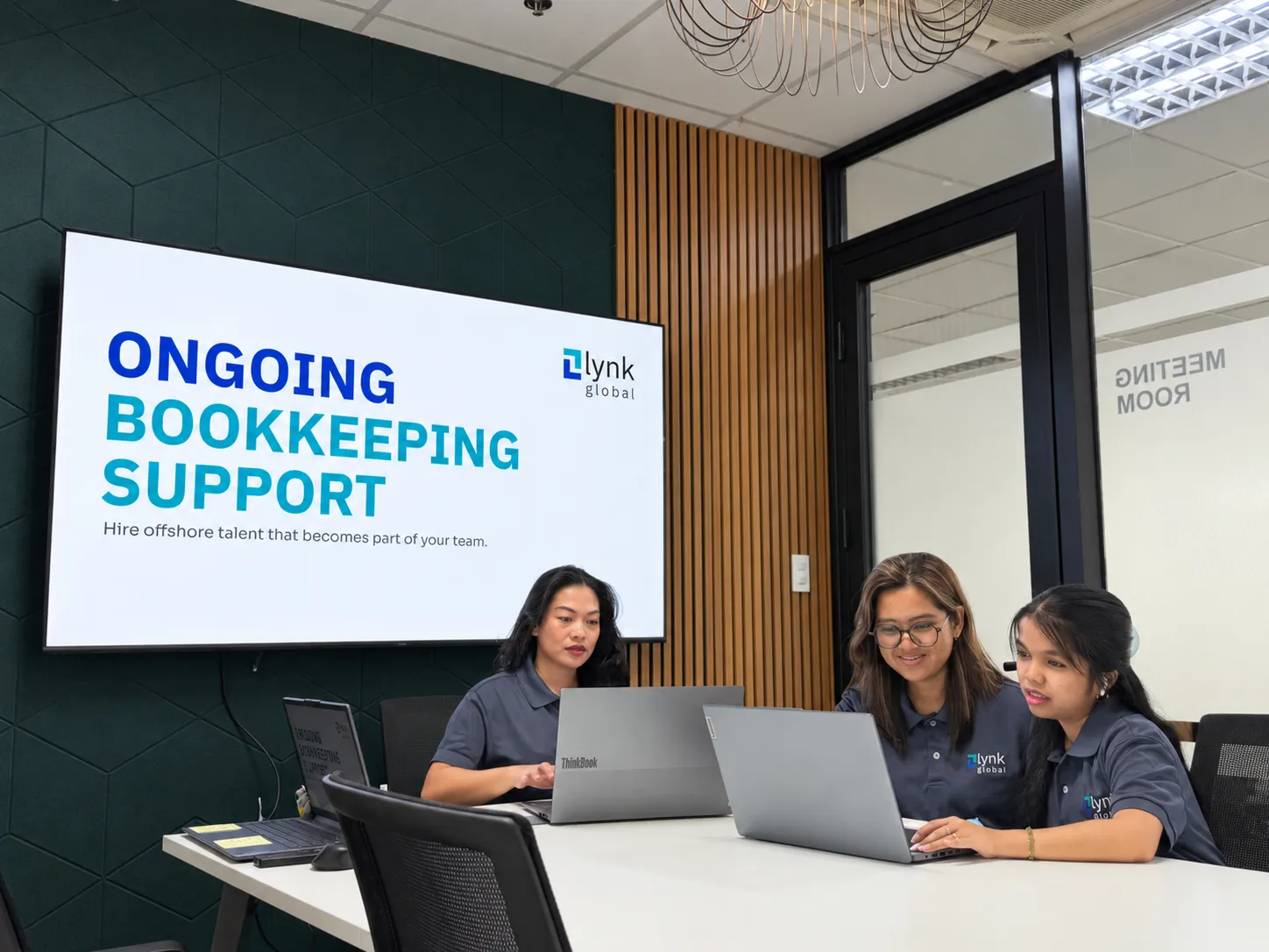 Three Lynk team members working on laptops in the Manila meeting room, with a bookkeeping support presentation on screen.