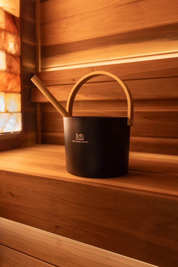 Black sauna bucket on cedar benches beside an illuminated salt wall