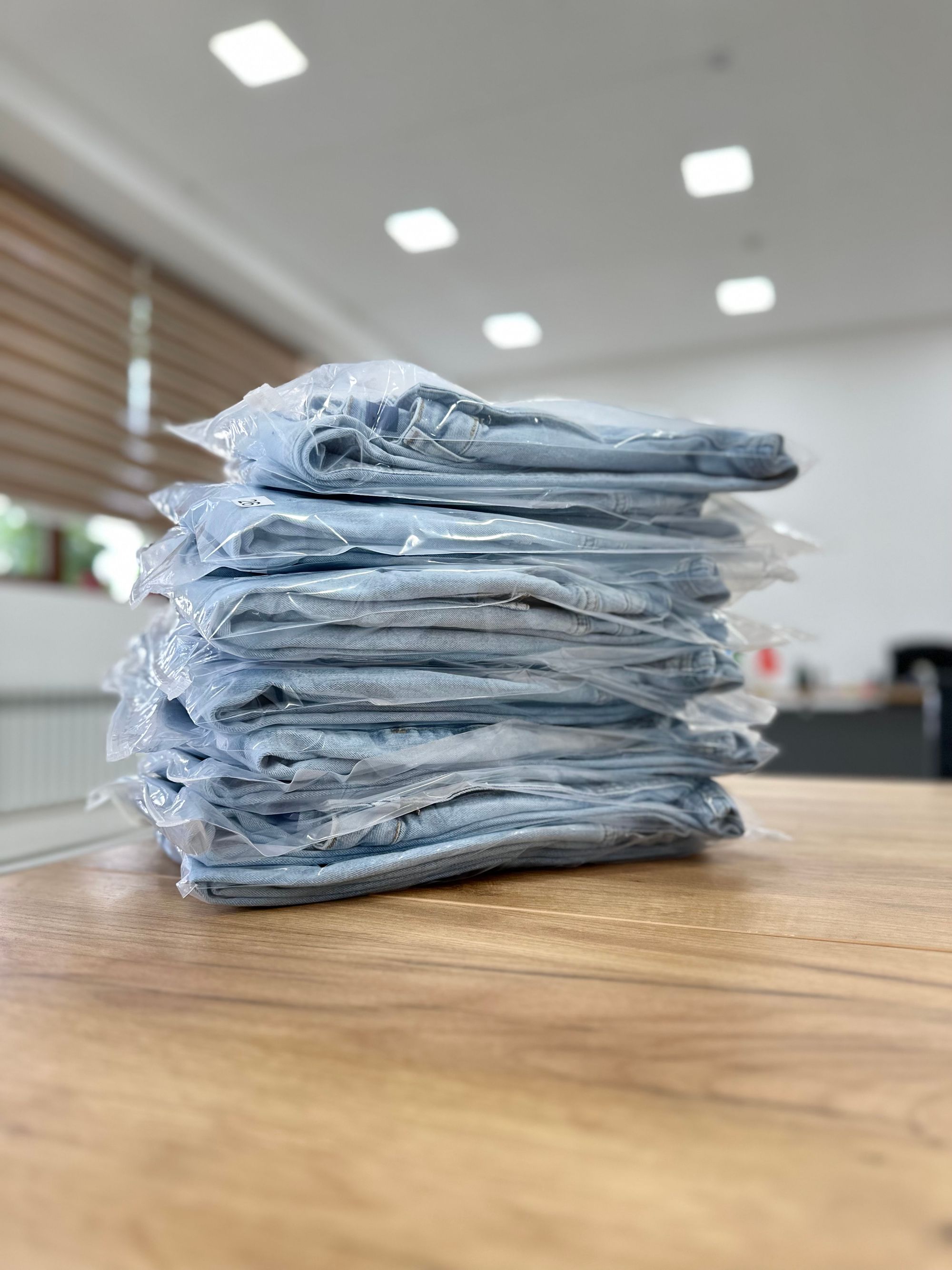 Gefaltete Jeans, jeweils einzeln in transparenten Beuteln verpackt, liegen als Stapel auf einem Holztisch.