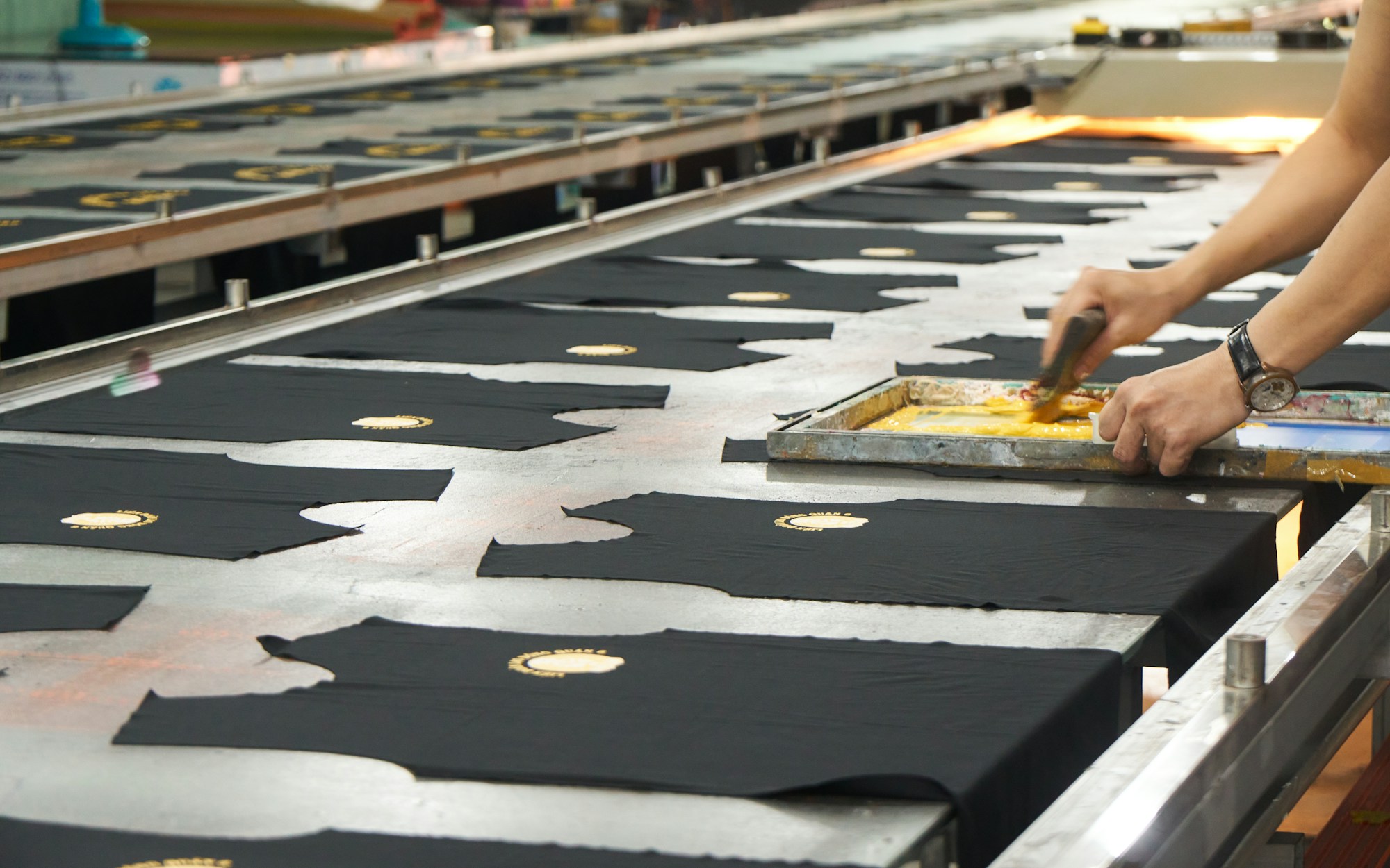 Hände bedrucken schwarze Bekleidungszuschnitte auf einem langen Drucktisch mit gelber Farbe im Siebdruckverfahren.