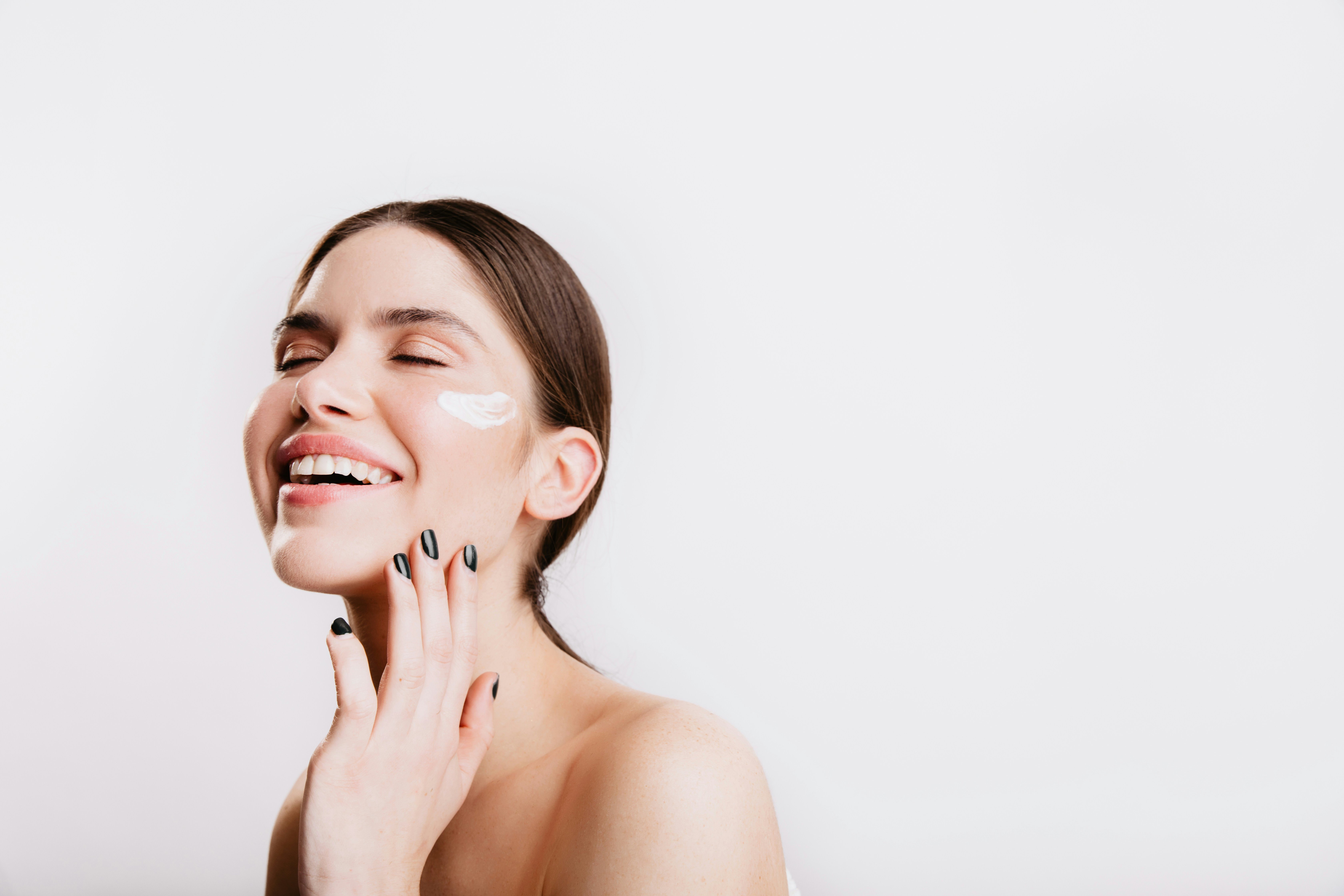 A woman laughing with her eyes closed and a dab of moisturizer on her cheek against a bright wall