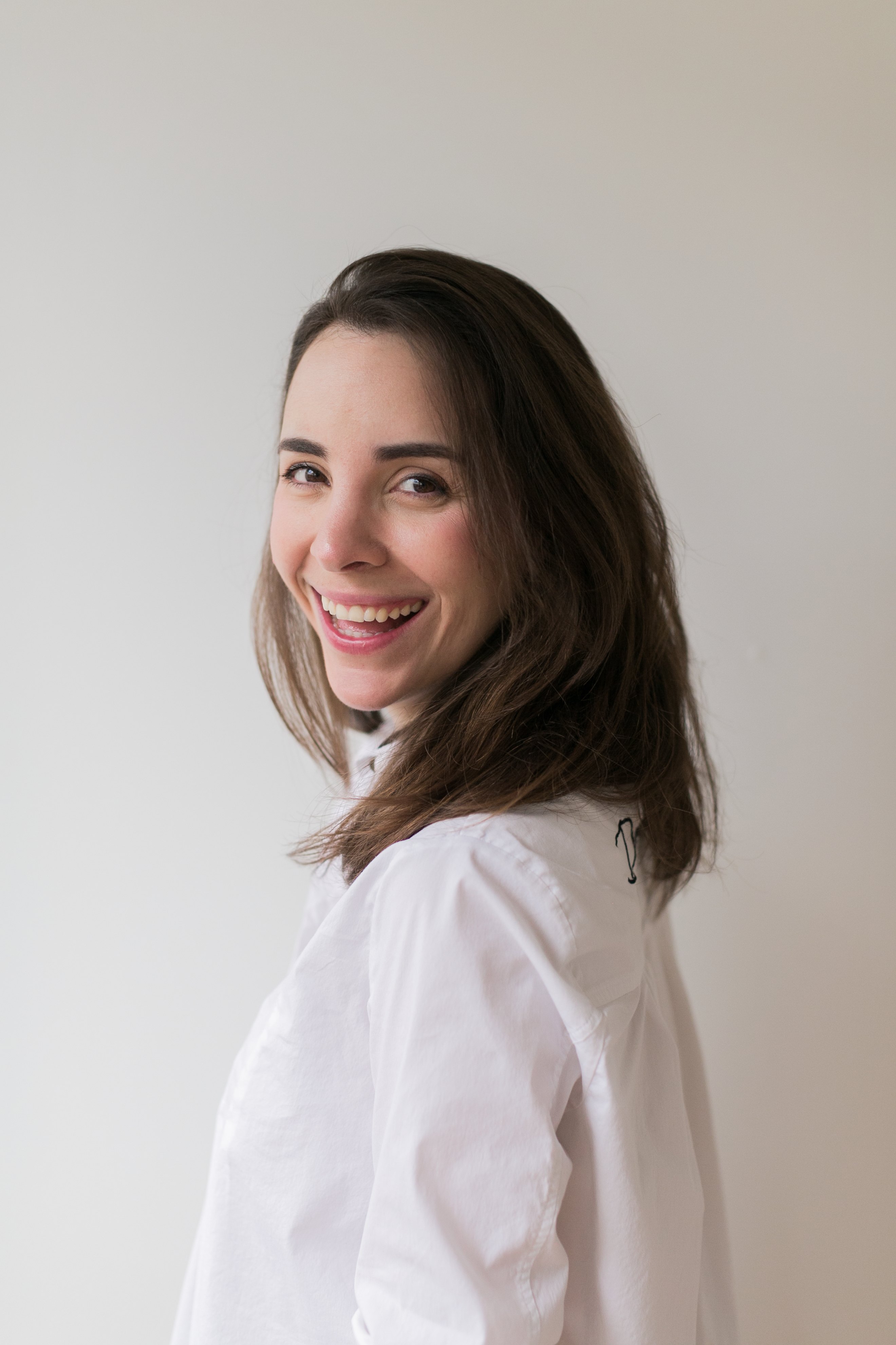 A smiling woman in a white shirt glancing back over her shoulder against a plain light wall