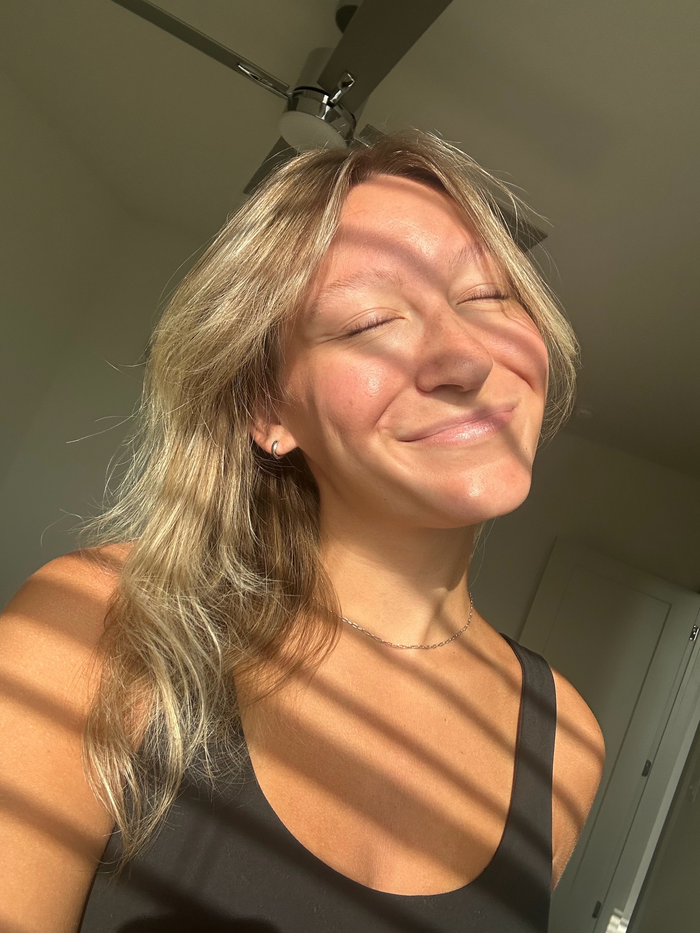 A smiling person with closed eyes in warm window light and shadows across their face