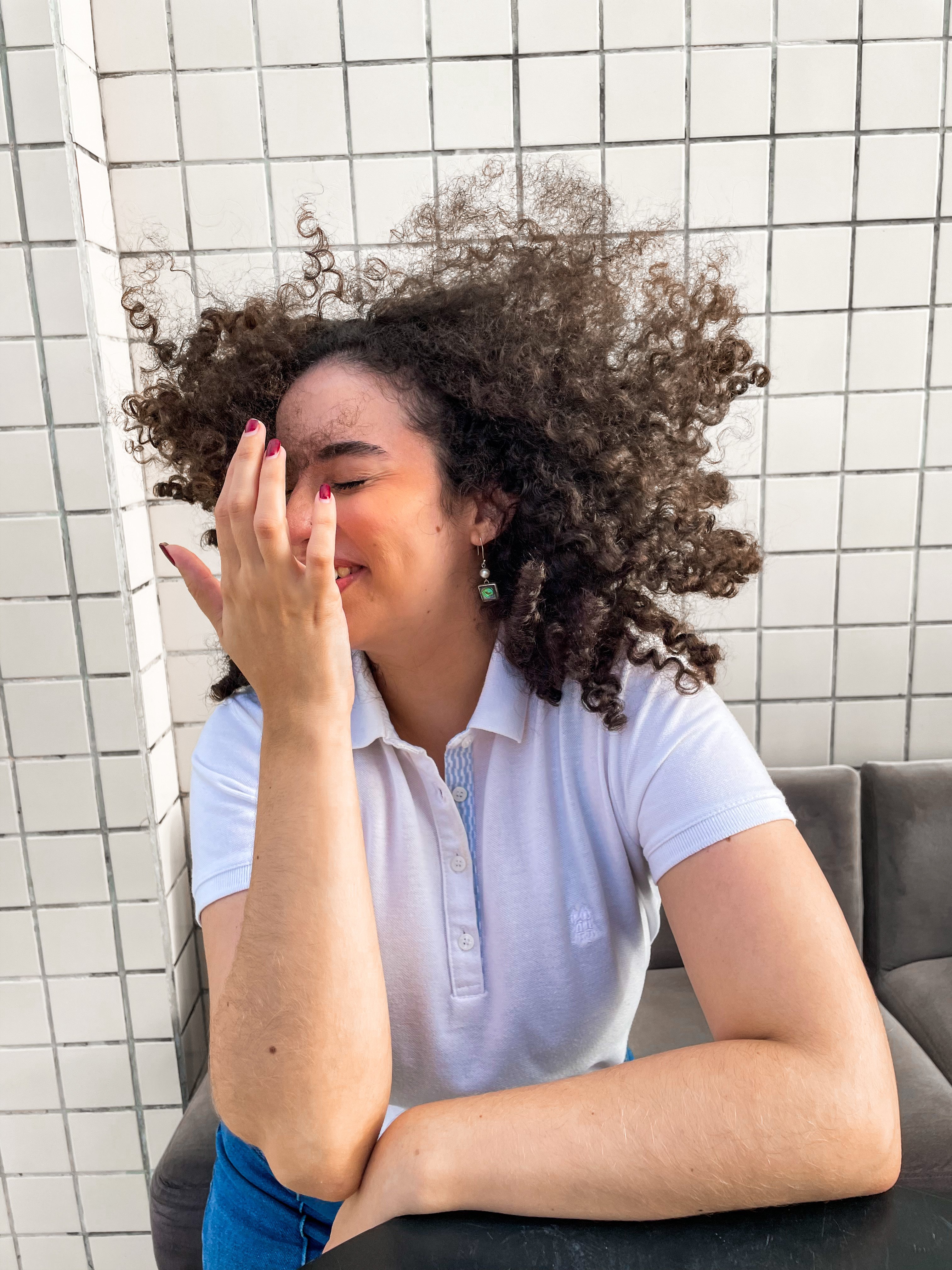 A person laughing with windswept curly hair, one hand raised to their face, seated against a white-tiled wall