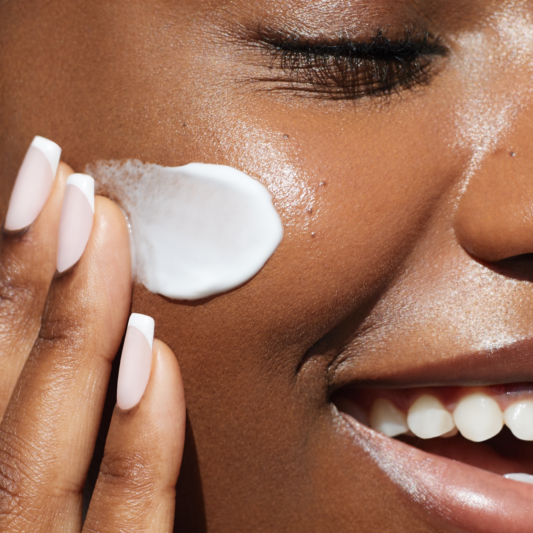 Close-up of a smiling woman with a smear of moisturizer on her cheek and her fingertips at the edge of the frame