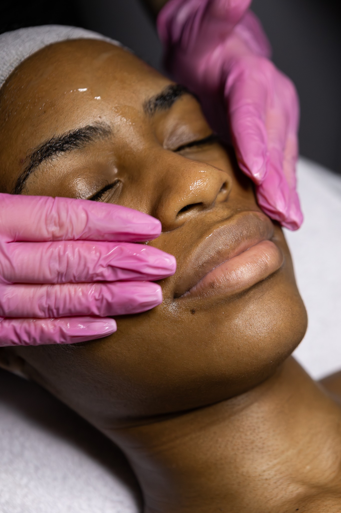 A client resting with eyes closed during a professional facial, with a provider's gloved hands supporting the face