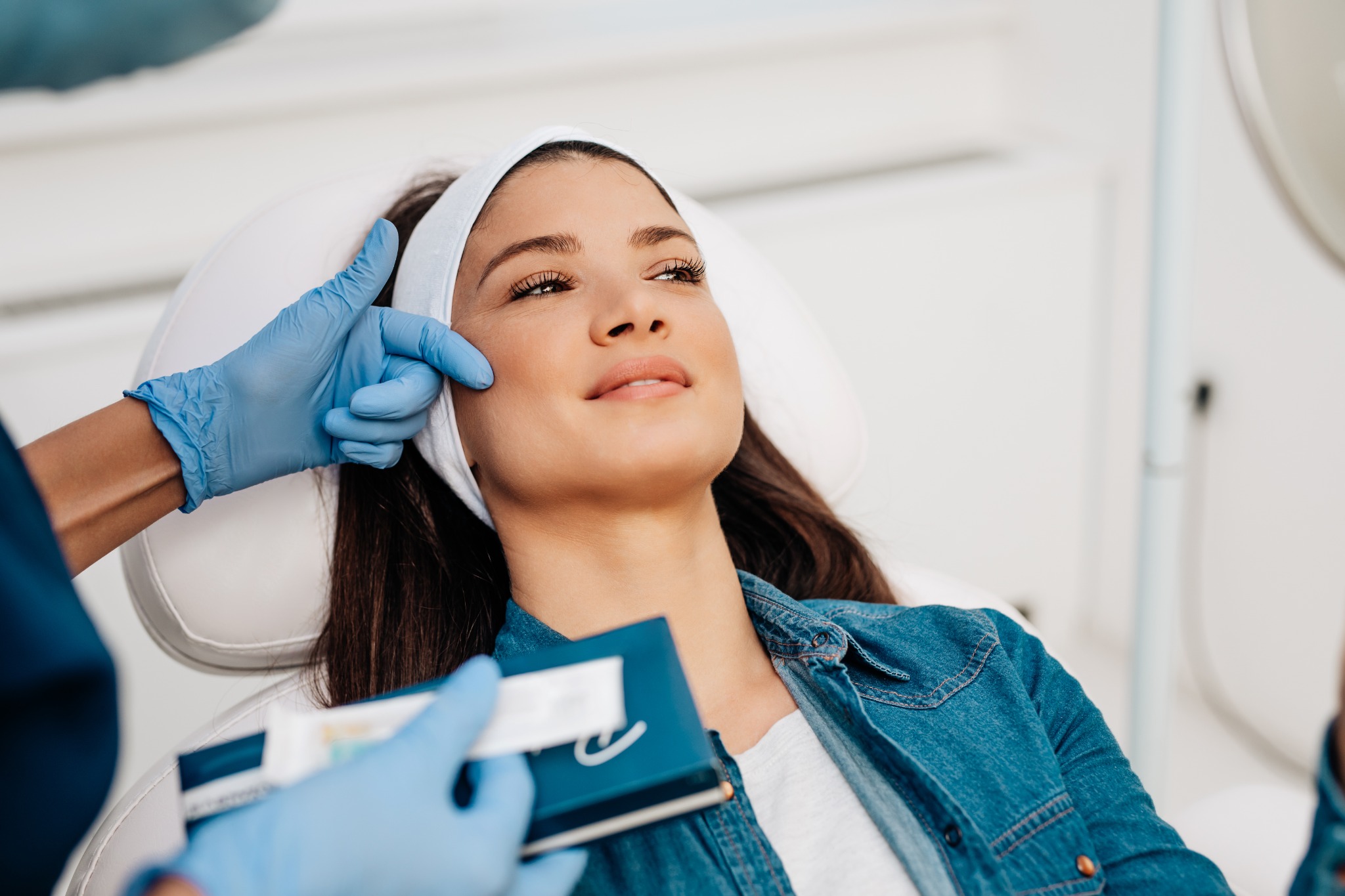 A relaxed person with a headband during a skin consultation while a gloved professional reviews their skin