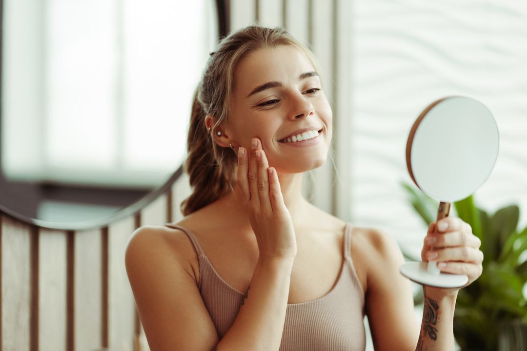 A smiling person touching their cheek while holding a hand mirror