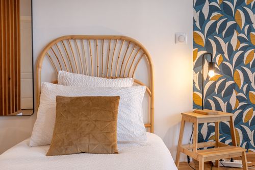 Detail of a single bed in La Chambrée with a rattan headboard and ochre cushion