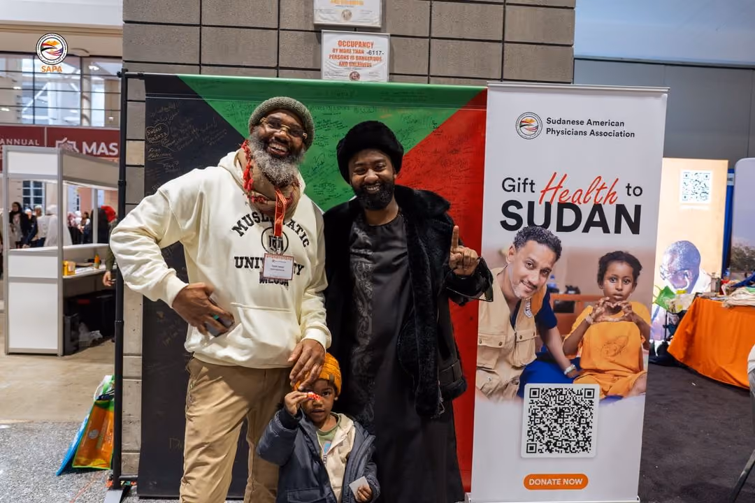 Two attendees in front of the signed photo backdrop and Gift Health to Sudan banner