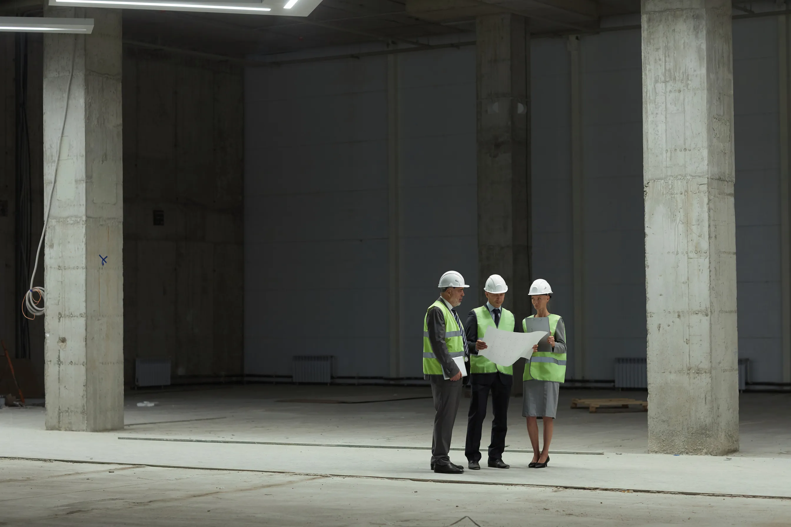 Three people wearing hard hats and high-visibility vests discuss a plan in an industrial hall.