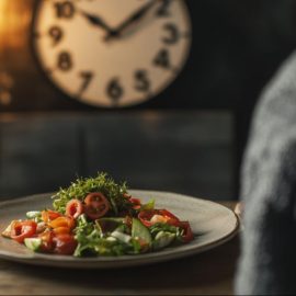 A plate of salad on a table with a clock on the wall in the background, representing intermittent fasting