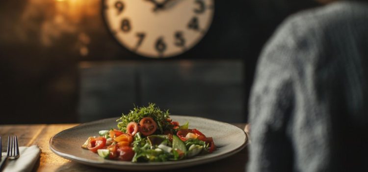 A plate of salad on a table with a clock on the wall in the background, representing intermittent fasting