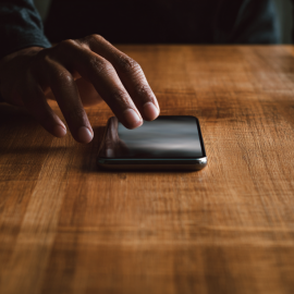 A hand reaching for a cell phone that's on a wooden table