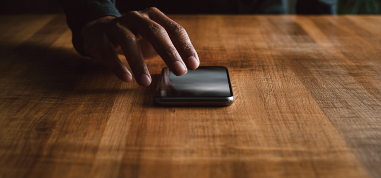 A hand reaching for a cell phone that's on a wooden table