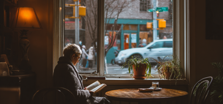 A woman with short hair and glasses sitting by a window in a city cafe and reading a book