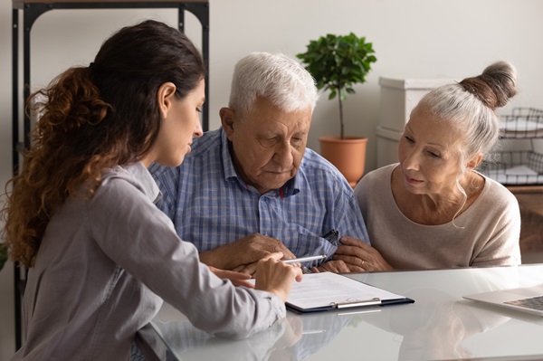 A female lawyer or consultant sitting at a desk and pointing at a document on a clipboard for an elderly couple, with the man looking intently at the paper.