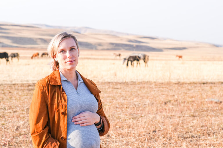 Caucasian pregnant woman touching her belly with horses grazing behind. Pregnancy journey in nature. Maternity experience