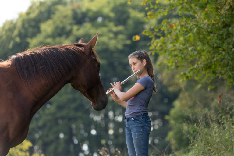 A girl playing a flute while standing in front of a horse.