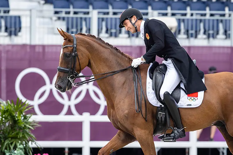 Man in dressage attire riding horse with Olympic saddle pad, leaning over and smiling