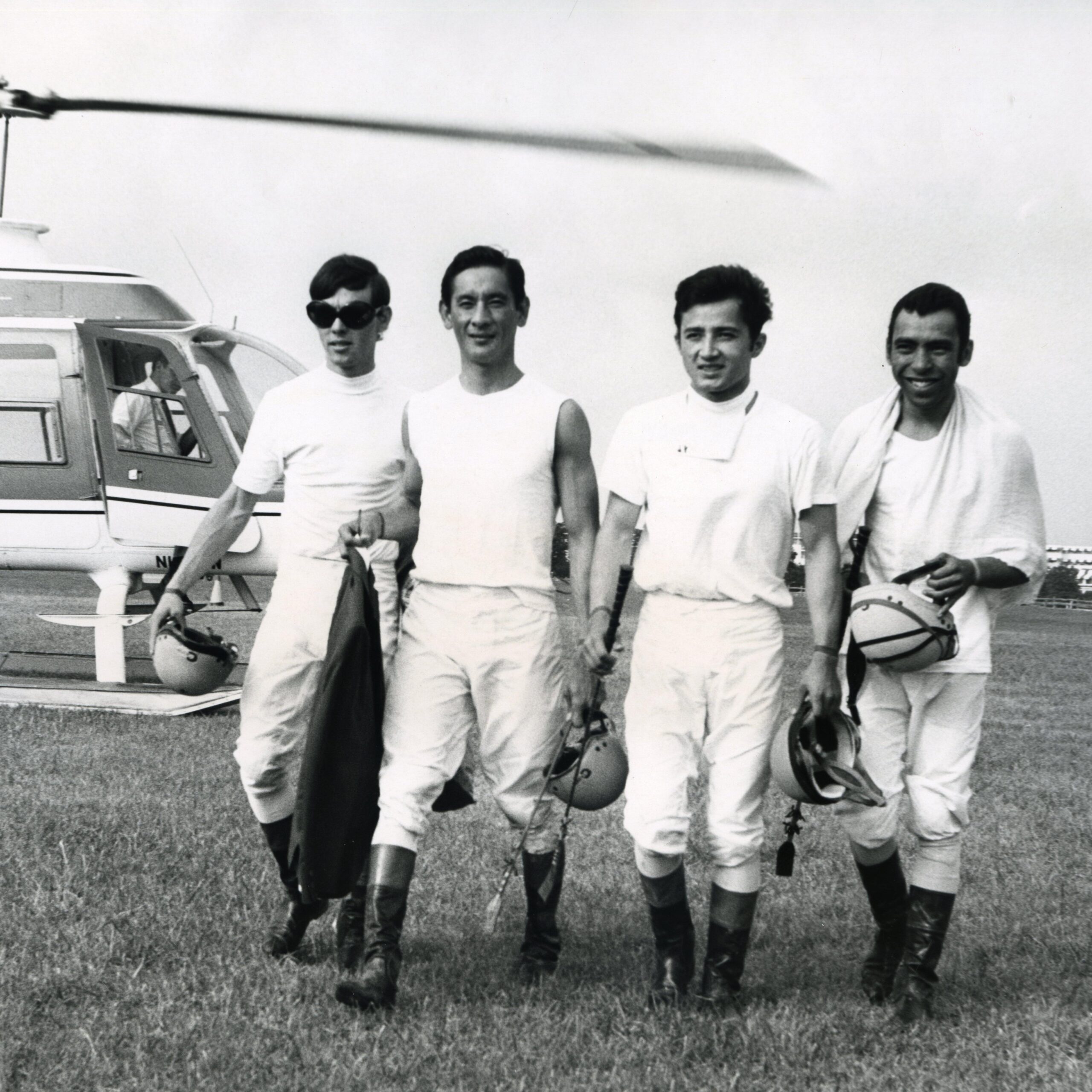 Black and white image of four men in jockeys' clothing walking together, helicopter in background
