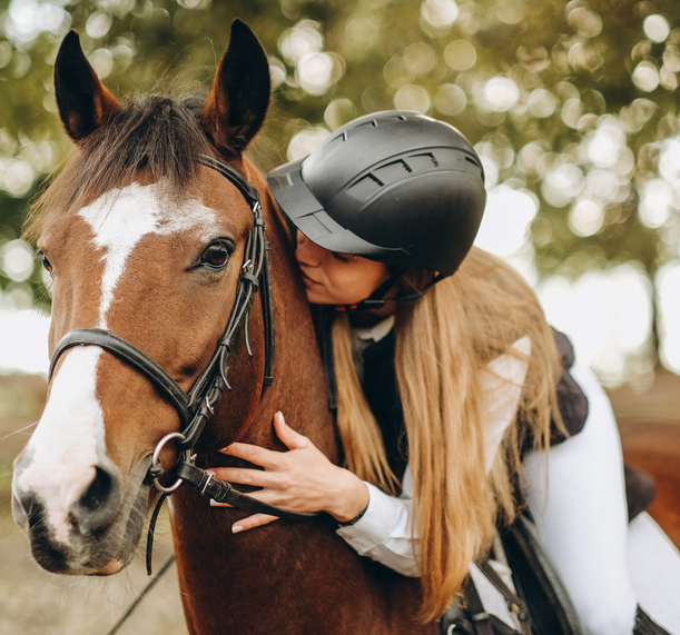 Closeup of helemted rider with long hair sitting on and hugging bridled bay horse with blaze
