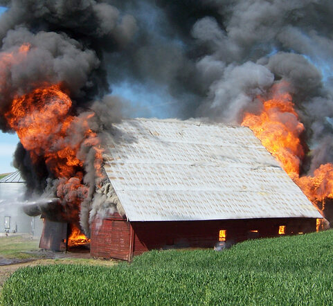 Red barn with flames erupting from both sides, grass in foreground and lots of smoke