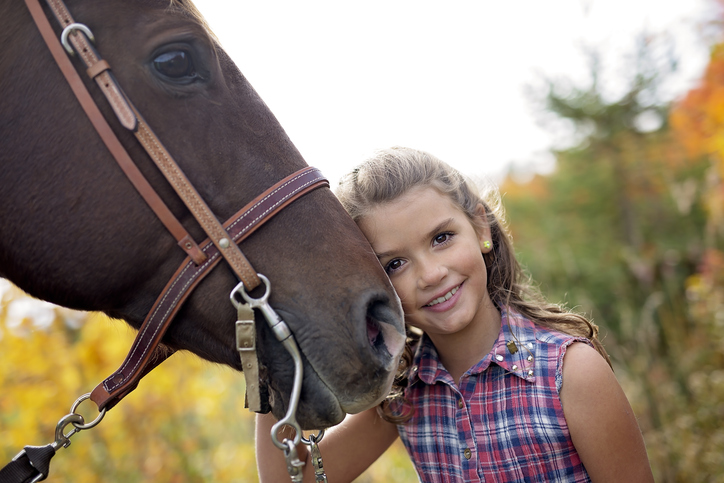 Young girl in tartan shit hugs bridled horse's nose while loking at the camera against autumn foliage backdrop
