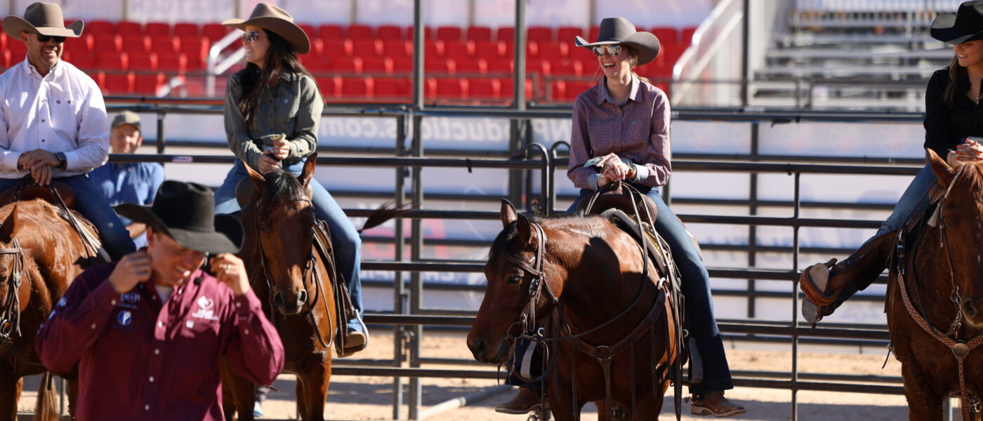 A group of riders in cowboy hats sit on horseback inside an outdoor arena. They appear relaxed and smiling, chatting with one another as they wait. A man in a maroon shirt walks in the foreground, slightly out of focus. The background shows red stadium seating, metal arena panels, and bright sunlight.