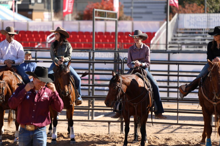 A group of riders in cowboy hats sit on horseback inside an outdoor arena. They appear relaxed and smiling, chatting with one another as they wait. A man in a maroon shirt walks in the foreground, slightly out of focus. The background shows red stadium seating, metal arena panels, and bright sunlight.