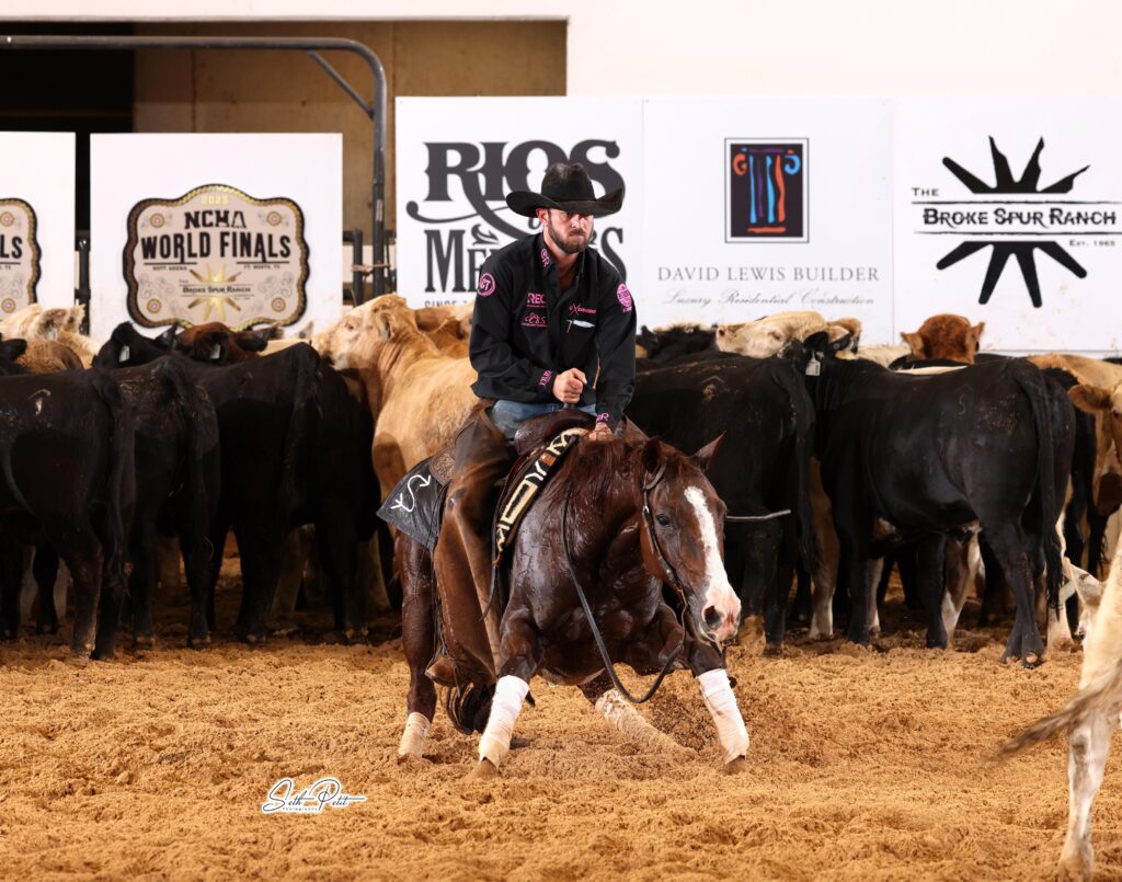 A rider in a black cowboy hat and show jacket guides a sorrel cutting horse as they work a cow away from the herd in an indoor arena. Large sponsor banners and cattle line the background, and sand kicks up under the horse’s hooves as it makes a sharp turn.