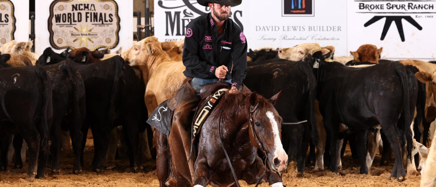 A rider in a black cowboy hat and show jacket guides a sorrel cutting horse as they work a cow away from the herd in an indoor arena. Large sponsor banners and cattle line the background, and sand kicks up under the horse’s hooves as it makes a sharp turn.