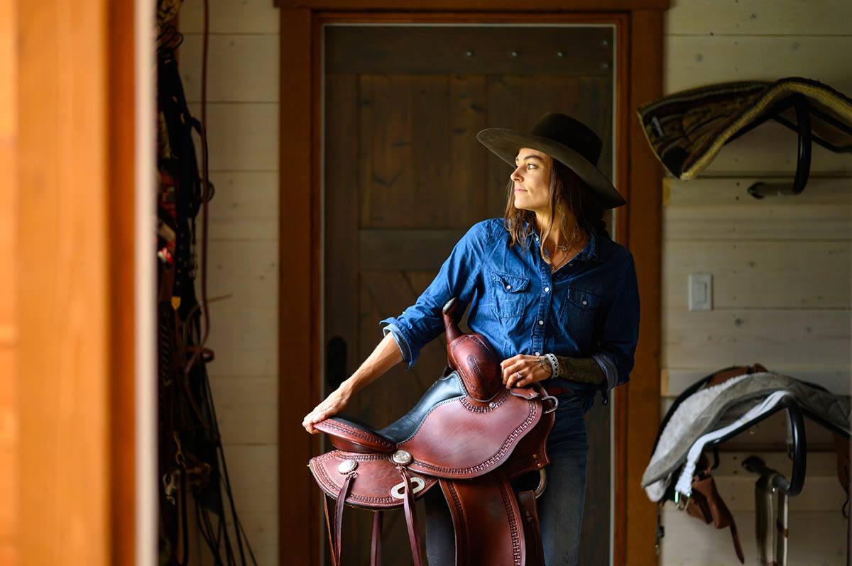 A woman carries a western saddle out of a tack room